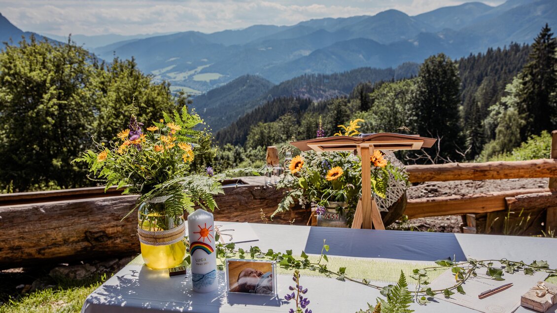 Ein Tisch mit Blumenarrangements und einem Getränk steht vor einem atemberaubenden Bergpanorama. Die Landschaft ist grün und die Sonne scheint. | © Manfred Müller