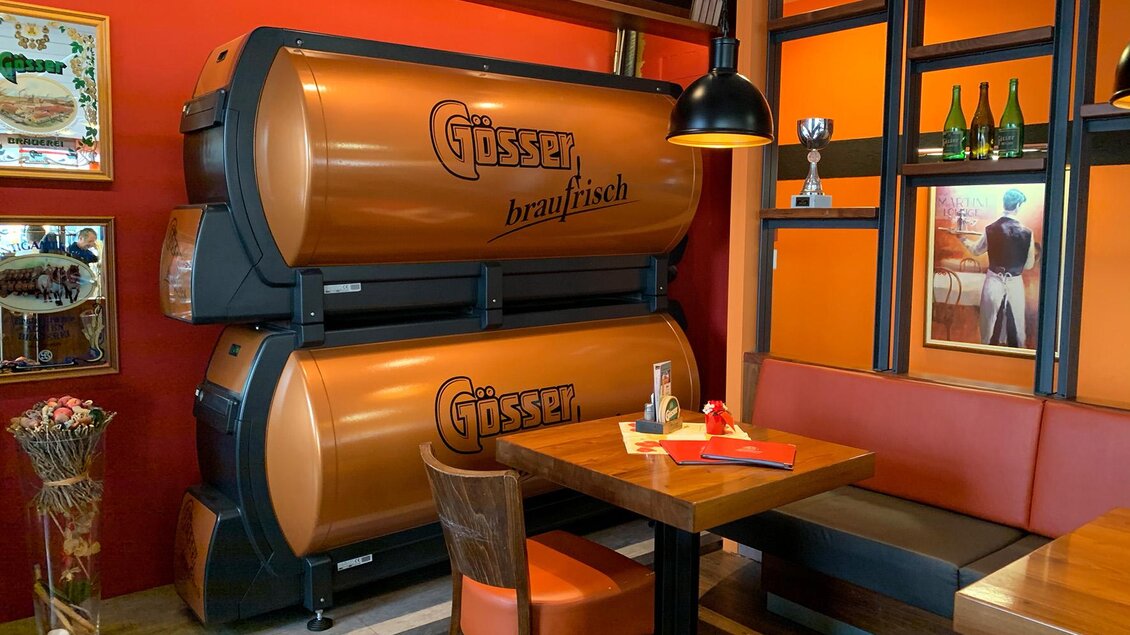 Eine gemütliche Sitzecke in einem Restaurant mit orangefarbenen Wänden. Im Hintergrund stehen große, orangefarbene Bierfässer von Gösser. | © M-Rast