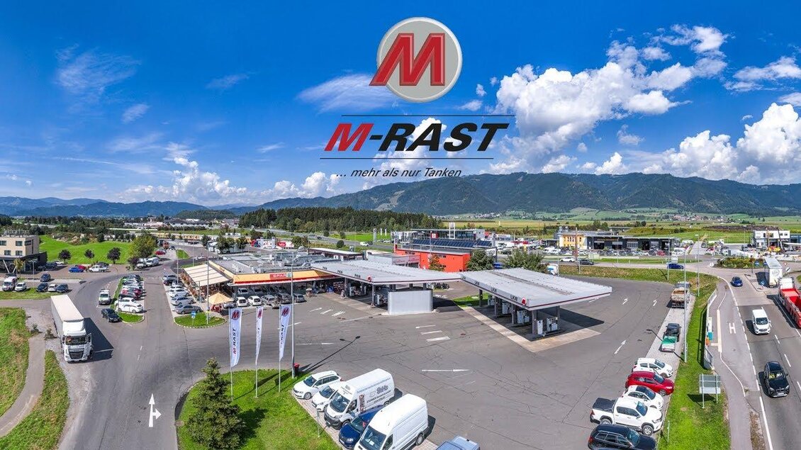 Ein modernes Rastangebot mit mehreren Gebäuden und einem Parkplatz. Im Hintergrund sind Berge und ein strahlend blauer Himmel sichtbar. | © M-Rast
