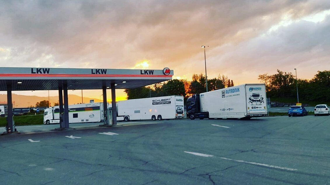 Ein Lkw-Parkplatz mit mehreren großen Lastwagen und Pkw. Der Himmel zeigt ein farbenprächtiges Sonnenuntergangslicht. | © M-Rast