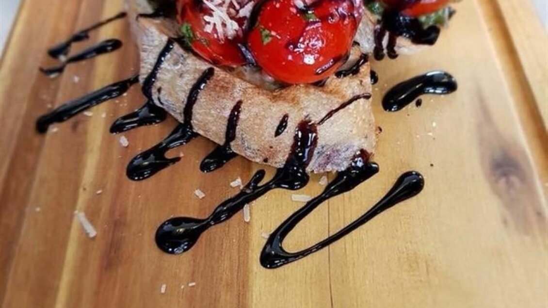 Ein köstliches Gericht auf einem Holzbrett. Es besteht aus geröstetem Brot, belegt mit Tomaten, Basilikum und einem Balsamico-Dressing. | © Stephan Friesinger