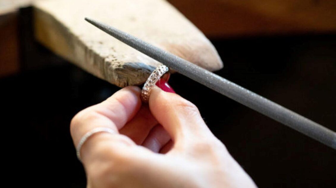 Eine Hand arbeitet an einem silbernen Ring mit einer Feile. Der Ring liegt auf einem Holzblock. | © Schmuck Design, Marion Buchsteiner