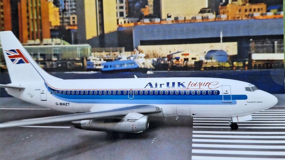 Ein Flugzeug von Air UK steht auf einer Landebahn vor einer modernen Stadtlandschaft. Die große Kulisse zeigt hohe Gebäude und Wasserfahrzeuge im Hintergrund. | © Helmut Kaiser