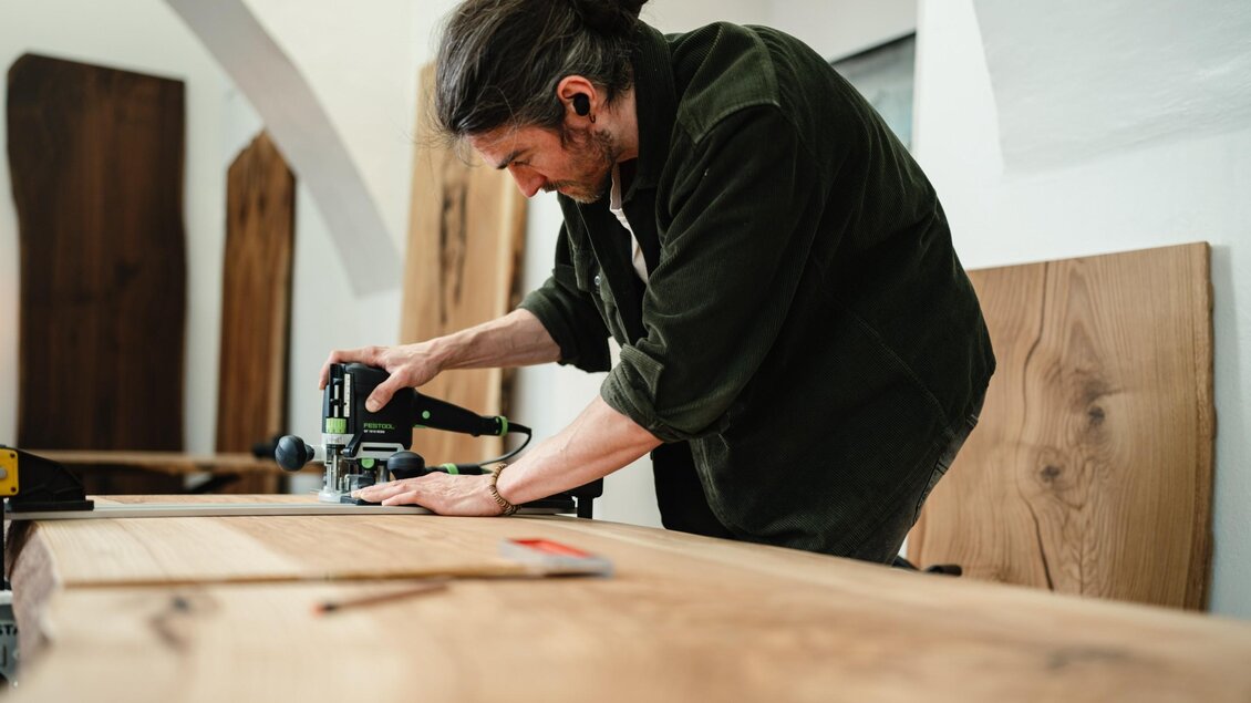 Manuel Stangl führt eine elektrische Oberfräse entlang einer Führungsschiene über eine Holzplatte im MORI Atelier. | © Bernd Grosseck