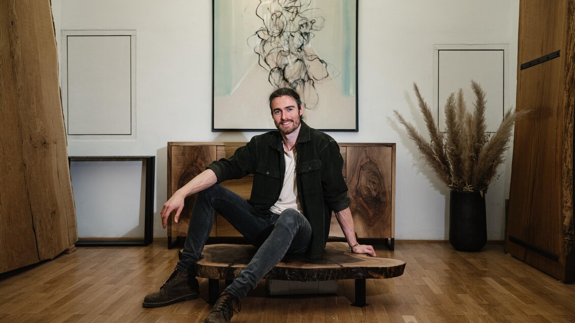Manuel Stangl sitzt auf einer Holzbank vor selbst gefertigten Möbeln aus Holz im MORI Atelier. | © Bernd Grosseck
