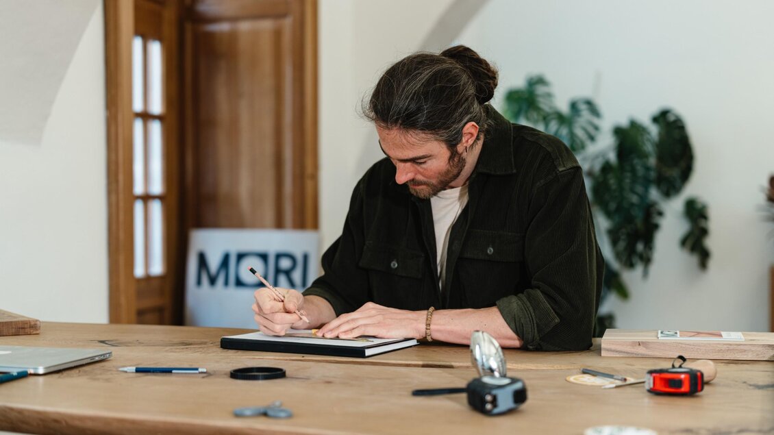 Manuel Stangl skizziert die Planung für ein neues Möbelstück mit Bleistift auf Papier im MORI Atelier. | © Bernd Grosseck