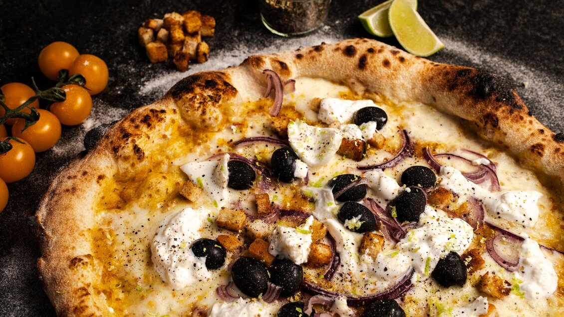Eine köstliche Pizza mit Mozzarella, schwarzen Oliven und Zwiebeln. Daneben liegen einige Cocktailtomaten und ein kleines Glas mit Gewürzen. | © Mundus kitchen