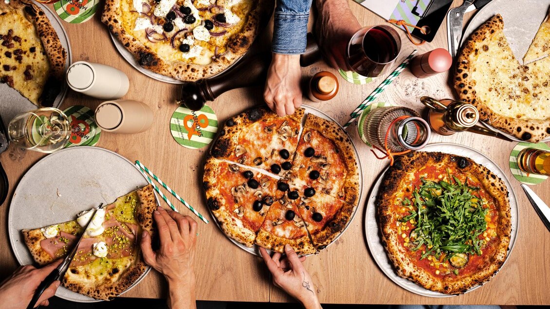 Ein Tisch voller köstlicher Pizzen und Getränke. Hände greifen nach dem Essen und genießen die gemeinsame Mahlzeit. | © Mundus kitchen