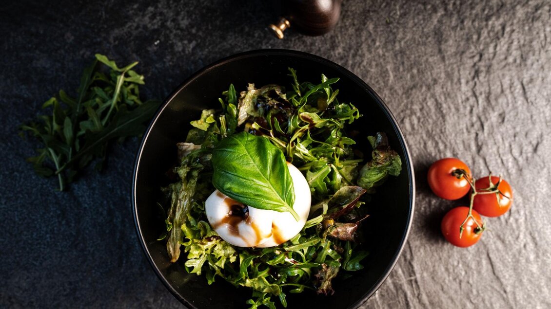 Ein elegantes Gericht mit frischem Salat und einer weißen Käsecreme in einer schwarzen Schüssel. Daneben liegen einige aromatische Kirschtomaten. | © Mundus kitchen