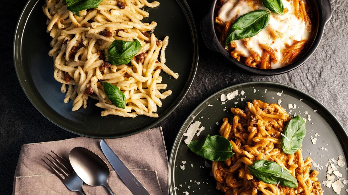 Zwei Teller Pasta, einer mit Basilikum und der andere mit Käse überbacken. Die Gerichte sind auf einem dunklen Tisch angerichtet. | © Mundus kitchen