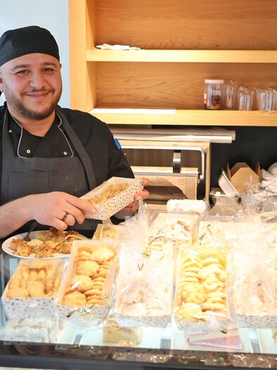 A laughing chef stands behind a table with various sweets. He is holding a treat in his hand and looks inviting. | © Stadt Bruck, Vassilev