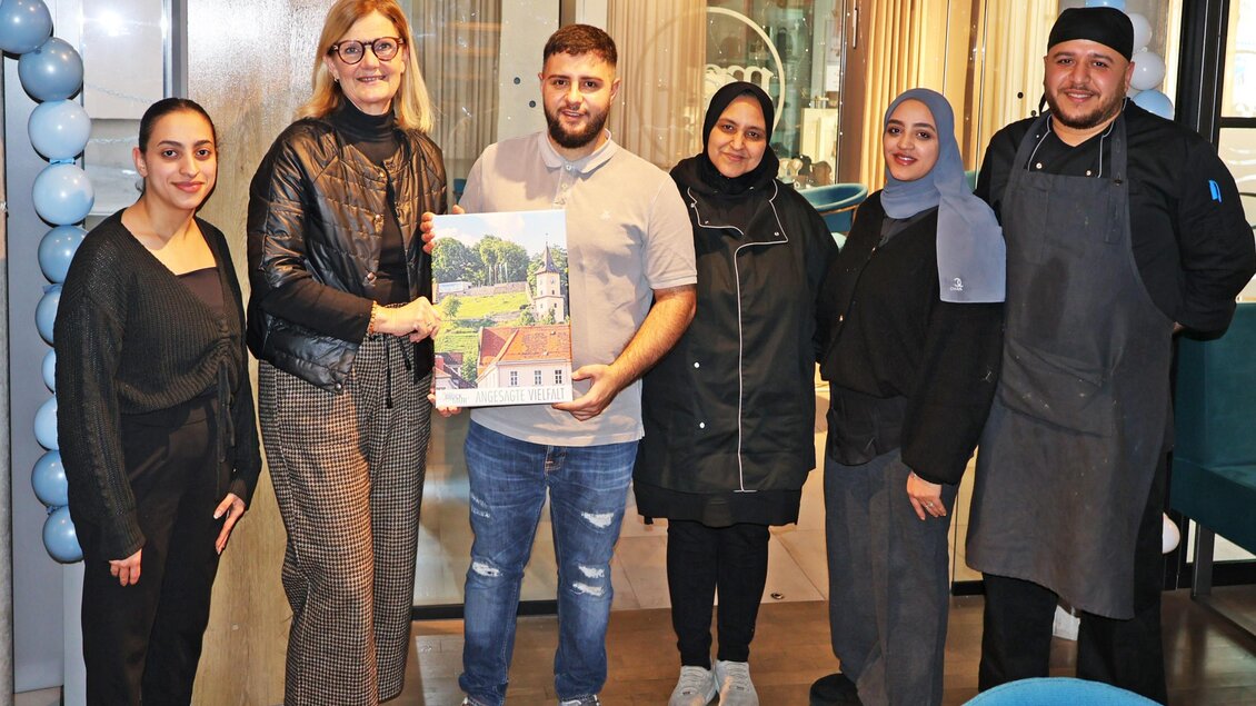 Eine Gruppe von sechs Personen steht zusammen in einem modernen Café. Sie lächeln und der Mann in der Mitte hält ein Plakat in der Hand. | © Stadt Bruck, Vassilev