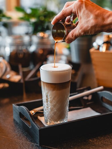 Eine Hand gießt einen Espresso in ein Glas mit schaumiger Milch. Der Tisch ist mit einem Serviertablett und weiteren Utensilien dekoriert. | © Thermen- & Vulkanland