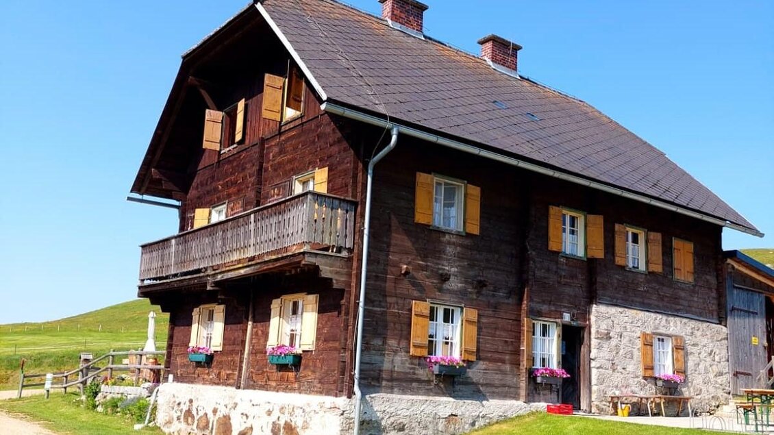 Ein traditionelles Holzhaus mit einem Balkon und bunten Fensterläden. Es steht auf einer Wiese unter blauem Himmel. | © Kaiser