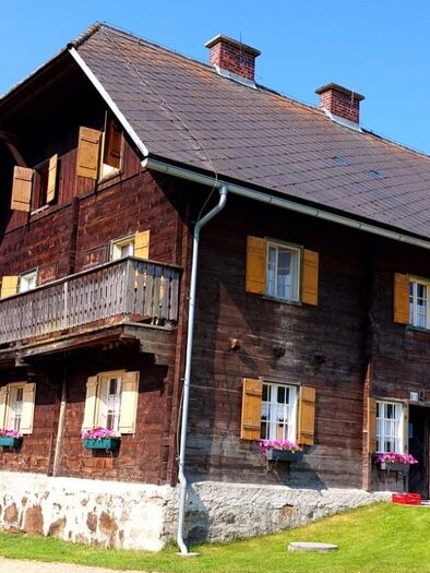 Ein traditionelles Holzhaus mit einem Balkon und bunten Fensterläden. Es steht auf einer Wiese unter blauem Himmel. | © Kaiser