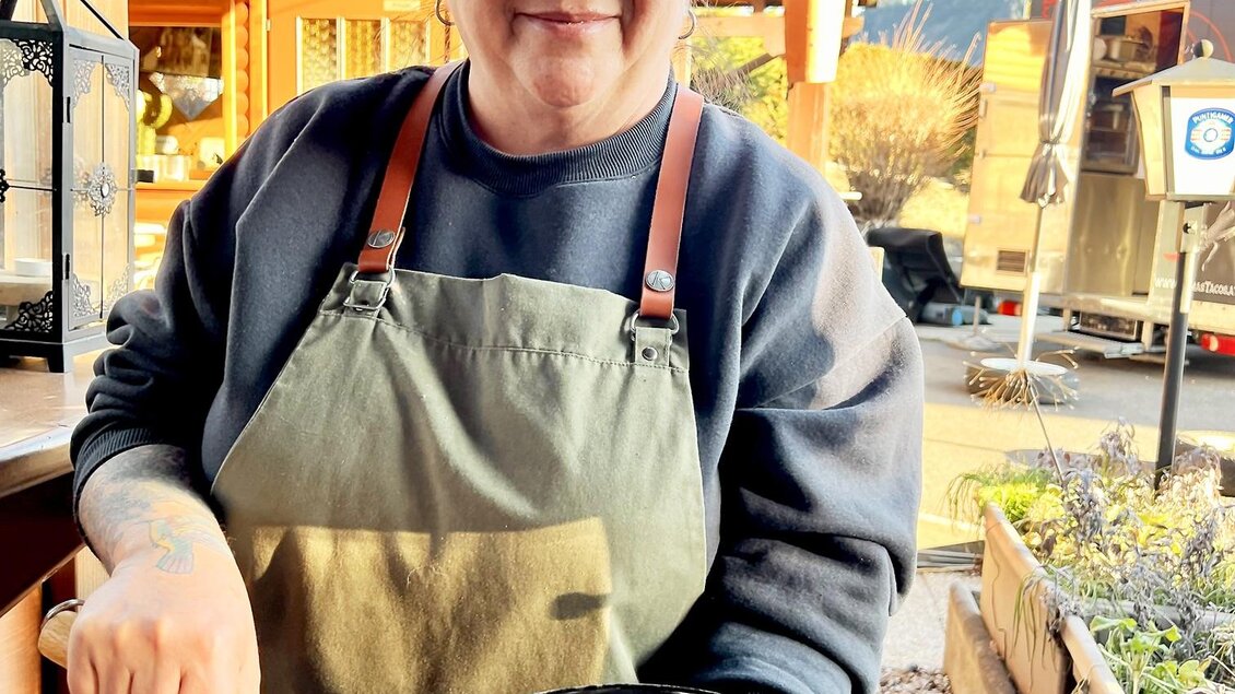 Eine fröhliche Frau in einem grünen Schürze hält eine Pfanne mit Essen. Im Hintergrund sind Holzbauten und die Natur zu sehen. | © Stadtmarketing Judenburg