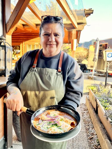 Eine fröhliche Frau in einem grünen Schürze hält eine Pfanne mit Essen. Im Hintergrund sind Holzbauten und die Natur zu sehen. | © Stadtmarketing Judenburg