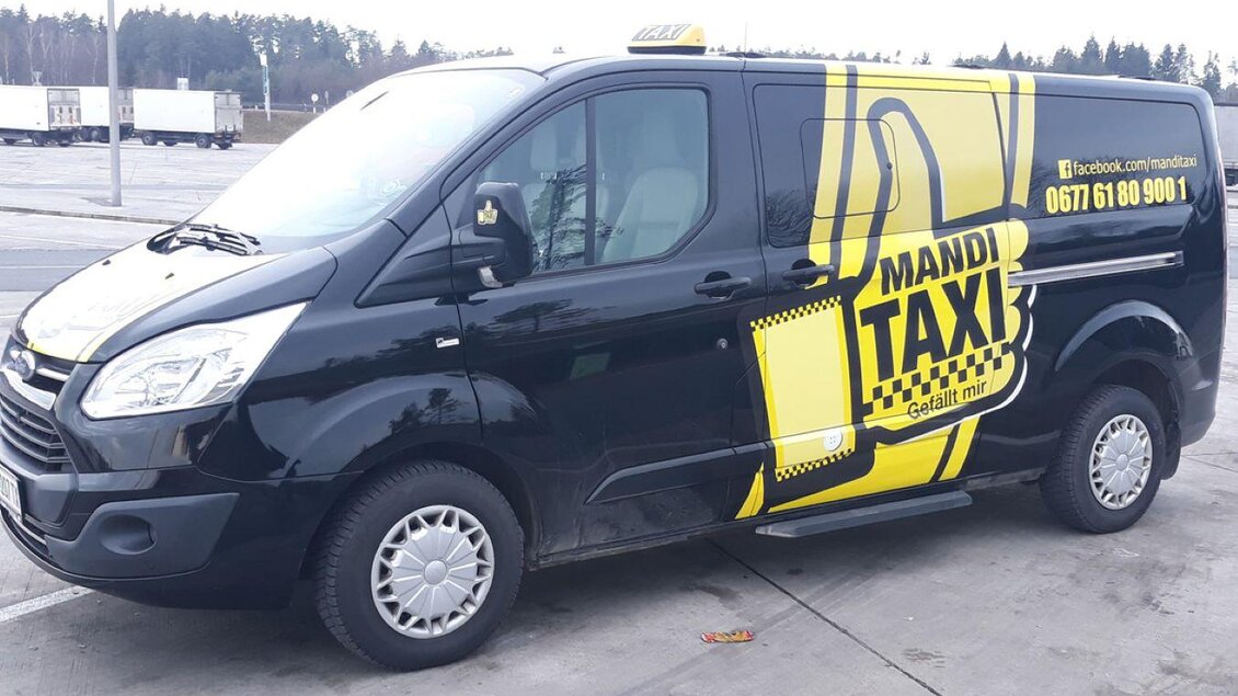 Ein schwarzer Van mit gelber Beschriftung und dem Logo eines Taxiunternehmens. Das Fahrzeug steht auf einem Parkplatz. | © Taxi Mandi und HandelsGmbH