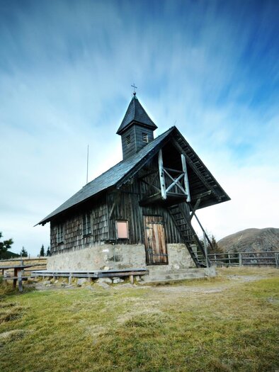 Eine alte Holzkirche auf einer Wiese mit sanften Hügeln im Hintergrund. Der Himmel ist blau mit bewegten Wolken. | © Eberhard