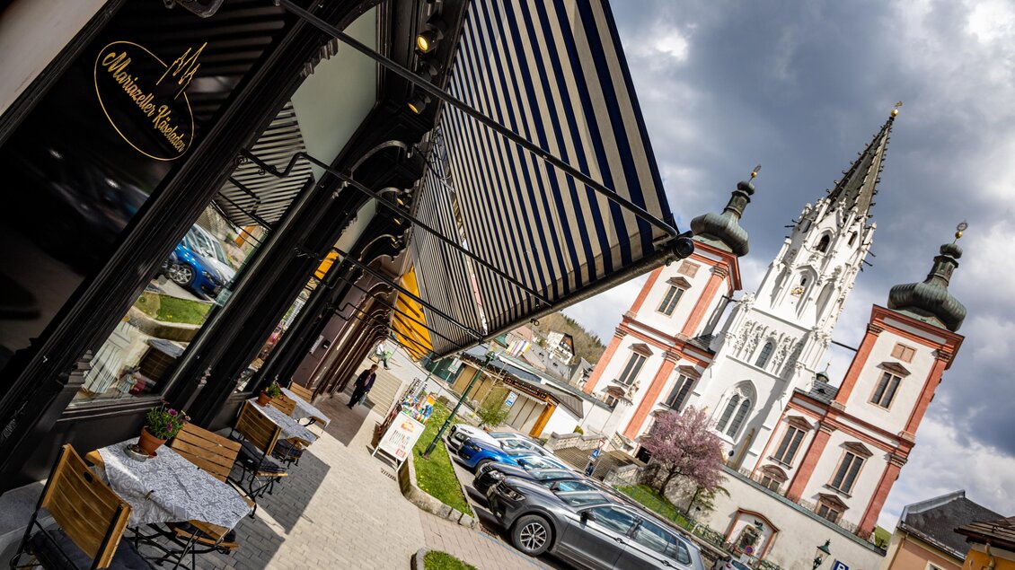 Ein malerischer Straßencafé mit Tischen im Freien, umgeben von historischen Gebäuden. Im Hintergrund erhebt sich eine beeindruckende Kirche mit einem hohem Turm.