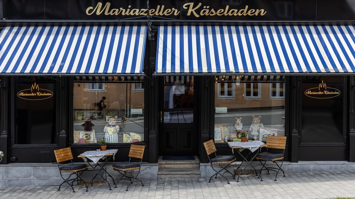Ein stilvoller Käseladen mit blauen Streifenmarkisen. Vor dem Laden stehen kleine Tische und Stühle auf dem Bürgersteig.