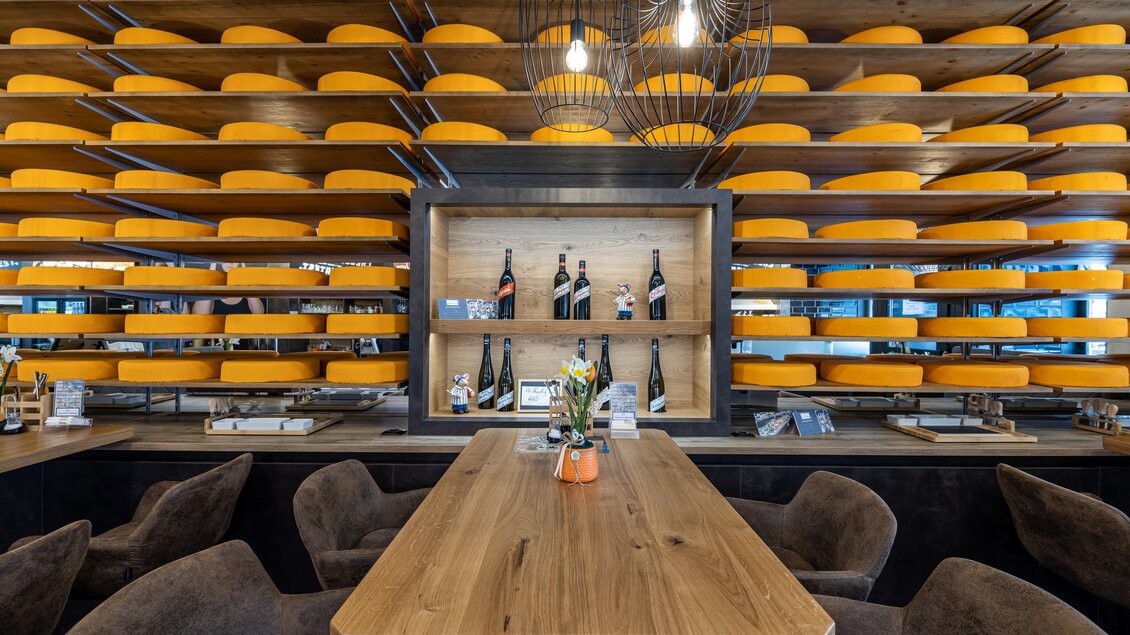 Ein stilvolles Restaurant mit einer holzfarbenen Tisch und gemütlichen Sesseln. Im Hintergrund ist eine Wand mit runden Käselaiben und dekorierten Flaschen zu sehen.