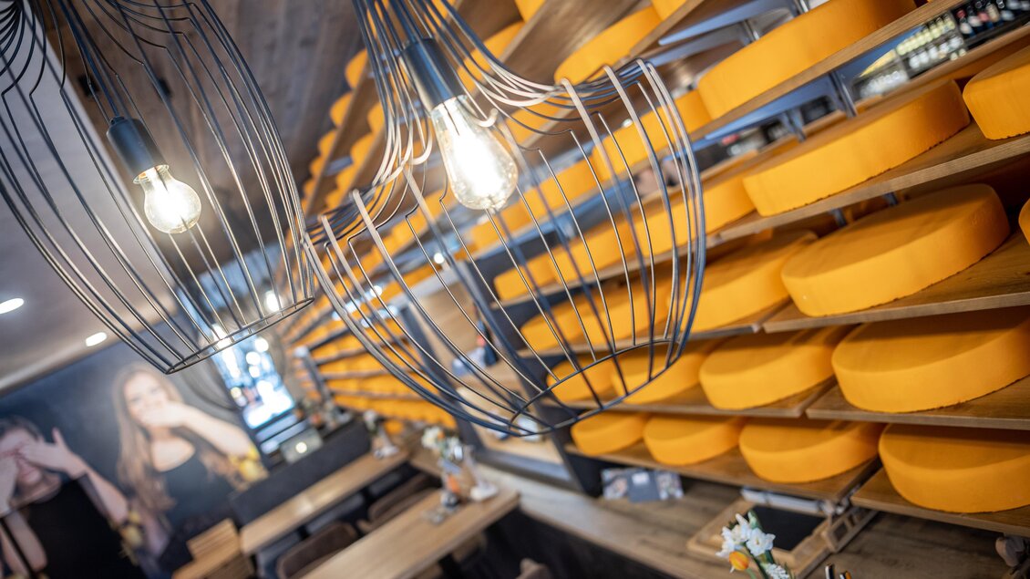 Ein gemütliches Restaurant mit hängenden Lichtern und Regalen voller orangefarbenem Käse. Die Einrichtung ist rustikal und einladend.