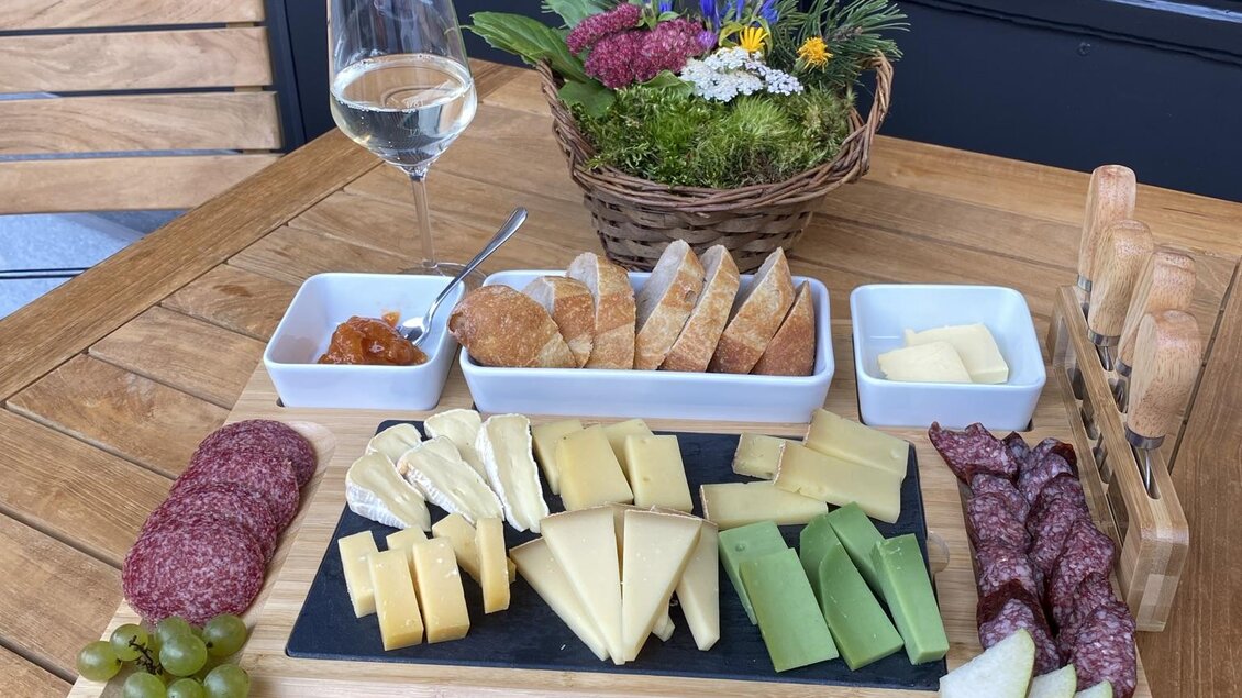 Ein schön angerichteter Tisch mit einer Käse- und Wurstplatte, ergänzt durch Trauben, Nüsse und Brot. Im Hintergrund steht ein Glas Weißwein und ein Blumengesteck.