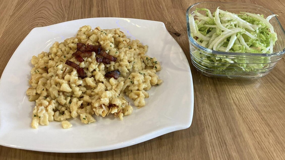 Ein Teller mit herzhaften Spätzle, garniert mit Speckstücken, liegt auf einem weißen Teller. Daneben befindet sich eine Schale mit frischem Krautsalat.