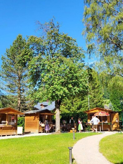<p>Ein malerischer Markt im Kurpark Altaussee mit vielen Holzständen umgeben von üppigem Grün. Die Sonne scheint hell auf das Markttreiben.</p> | © Petra Kirchschlager