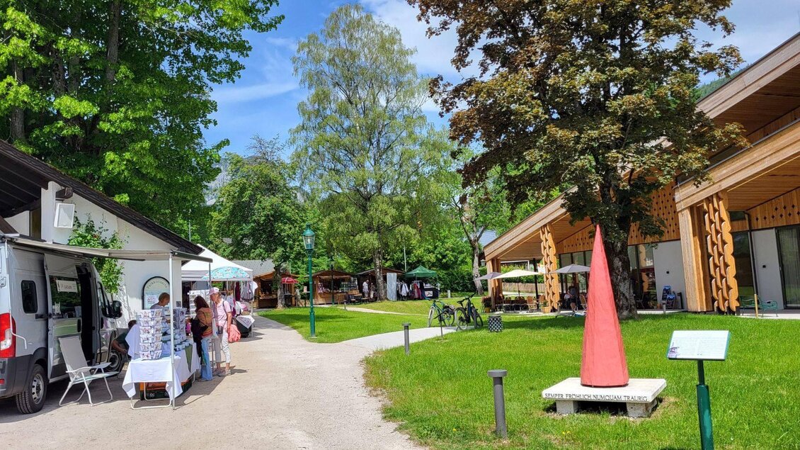 Ein lebendiger Außenbereich mit Ständen und einem roten Kunstwerk. Umgeben von Bäumen und einem klaren Himmel. | © Petra Kirchschlager