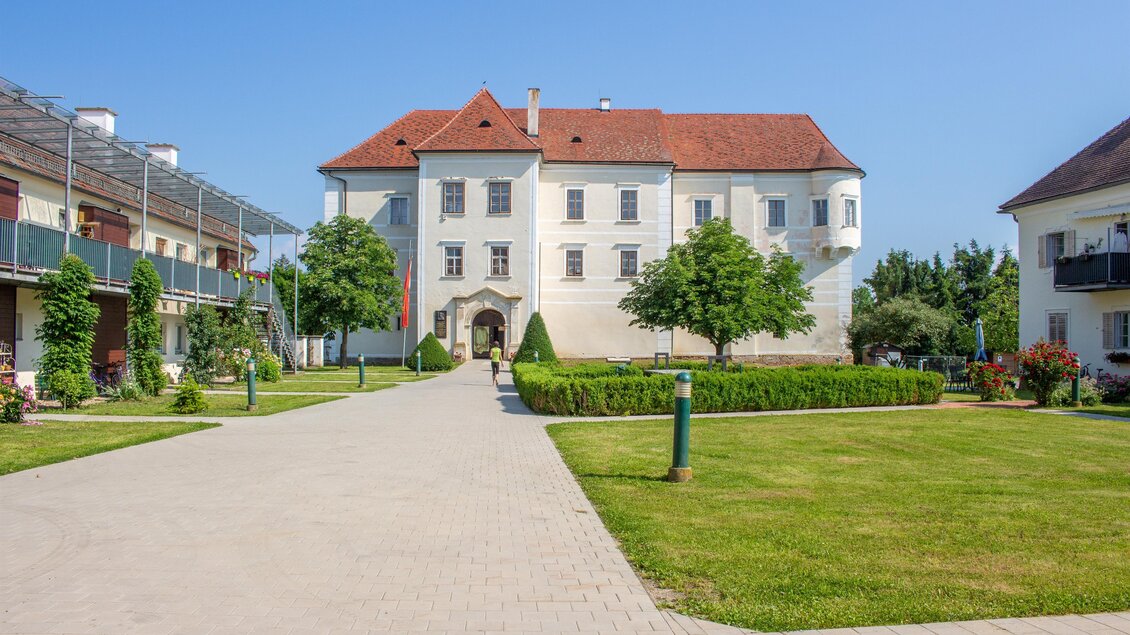 Ein schönes Gebäude mit einem roten Dach, umgeben von gepflegten Grünflächen und Bäumen. Der klare Himmel und die sonnige Atmosphäre schaffen eine einladende Stimmung. | © Sandra Brünner