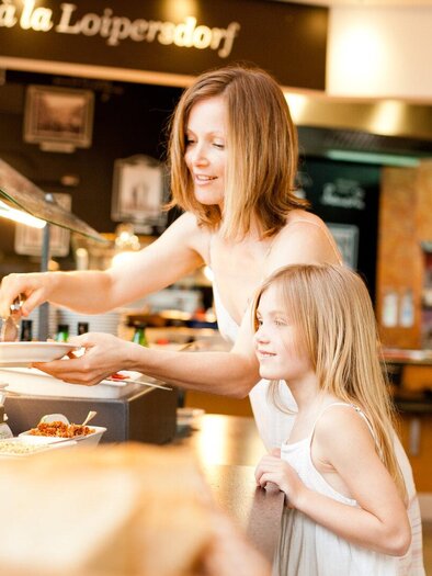 <p>Eine Frau und ein Mädchen stehen an einem Buffet und wählen Essen aus. Im Hintergrund sind frische Speisen und eine einladende Umgebung zu sehen. Marktrestaurant Thermenresort Loipersdorf in Bad Loipersdorf.</p> | © Thermenresort Loipersdorf