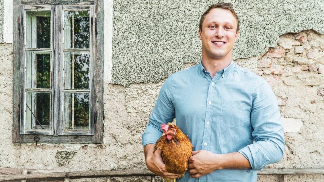 Ein junger Mann steht vor einer alten Wand und hält ein Huhn in den Händen. Hinter ihm ist ein Fenster mit Holzrahmen zu sehen. | © Martin´s das schmeckt