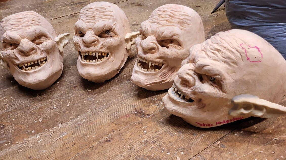 Vier groteske Masken mit verzerrten Gesichtern und sichtbaren Zähnen stehen auf einer Holzoberfläche. Die Masken sind in einer handwerklichen Werkstatt platziert. | © Rene Pörr