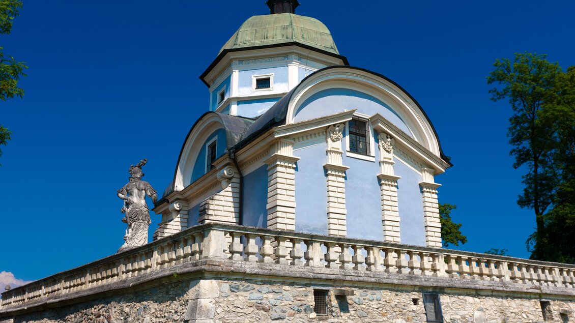 Ein schmuckes Gebäude mit einer grünen Kuppel und einer eleganten Fassade. Es steht auf einer Steinmauer, umgeben von Bäumen und klarem blauem Himmel. | © SYMBOL | SCHIFFER