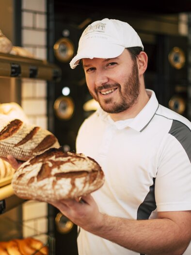 A baker smiles warmly and holds two freshly baked loaves of bread in his hands. In the background, various breads and rolls can be seen on a display shelf. | © Tourismusverband Feldbach/ B. Bergmann
