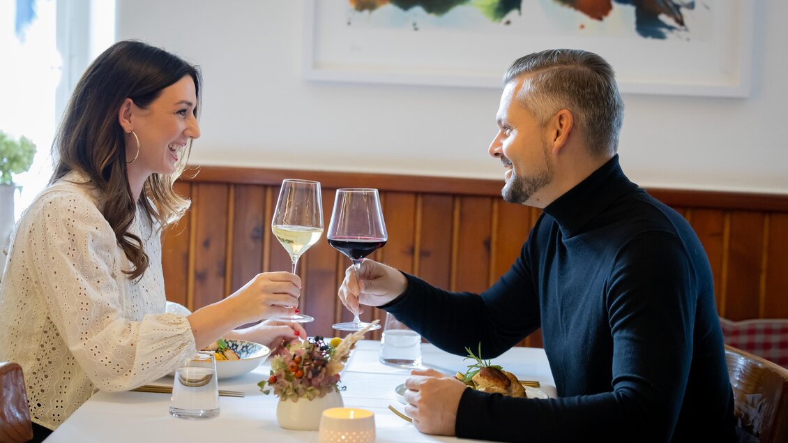 Ein Paar sitzt an einem Tisch in einem Restaurant und hebt Gläser zur traditionellen Anstoßen. Sie lächeln sich an und genießen ihr Essen in einer gemütlichen Atmosphäre. | © Stadthotel zur goldenen Krone