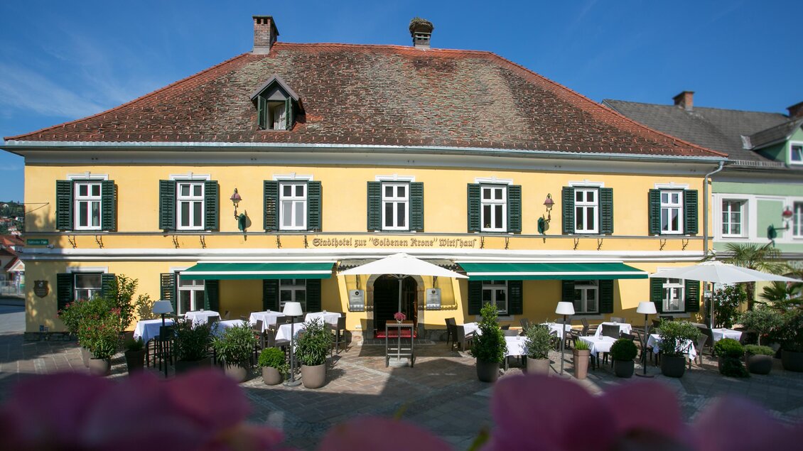 Ein charmantes gelbes Gebäude mit einer Terrasse und Sonnenschirmen. Blumen und Grünpflanzen umgeben den Eingangsbereich. | © Stadthotel zur goldenen Krone