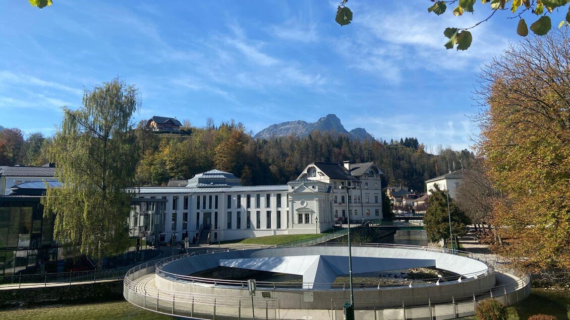 Die über dem Zusammenfluss der Grundlseer und Altausseer Traun schwebende Mercedessternbrücke, umgeben von Bäumen und Bergen des Ausseerlandes. Dahinter befindet sich das schöne Gebäude des alten Kurmittelhauses in Bad Aussee. Der Himmel ist klar und blau.
 | © TVB Ausseerland Salzkammergut/ Jana Grill