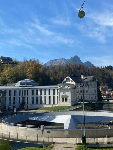 Die über dem Zusammenfluss der Grundlseer und Altausseer Traun schwebende Mercedessternbrücke, umgeben von Bäumen und Bergen des Ausseerlandes. Dahinter befindet sich das schöne Gebäude des alten Kurmittelhauses in Bad Aussee. Der Himmel ist klar und blau.
 | © TVB Ausseerland Salzkammergut/ Jana Grill