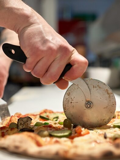 A pizza maker is cutting a fresh pizza with a pizza cutter. The pizza is topped with vegetables and looks delicious. | © AdobeStock_152717580
