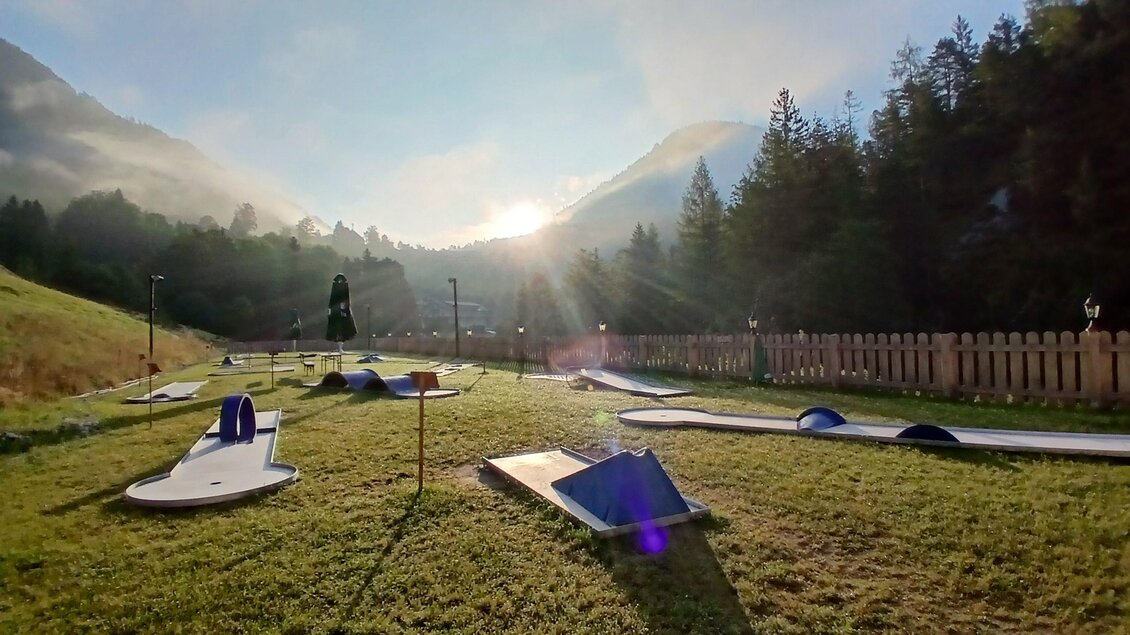 Eine sonnige Landschaft mit einem Minigolfplatz und Bergen im Hintergrund. Die Umgebung ist grün und friedlich, ideal für Freizeitaktivitäten.