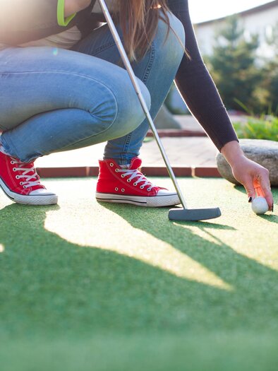 Eine Person spielt Minigolf und beugt sich über den Putt. Es ist sonnig und die Umgebung ist grün. | © AdobeStock_70570315 (1)_Minigolf