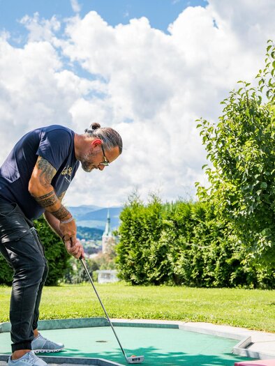 <p>Person spielt Minigolf im Freien in der Lipizzanerheimat in Voitsberg. Der Spieler ist in einer sportlichen Pose und zielt auf das Loch, während im Hintergrund eine grüne Landschaft und Bäume zu sehen sind.</p> | © Region Graz - Mias Photoart