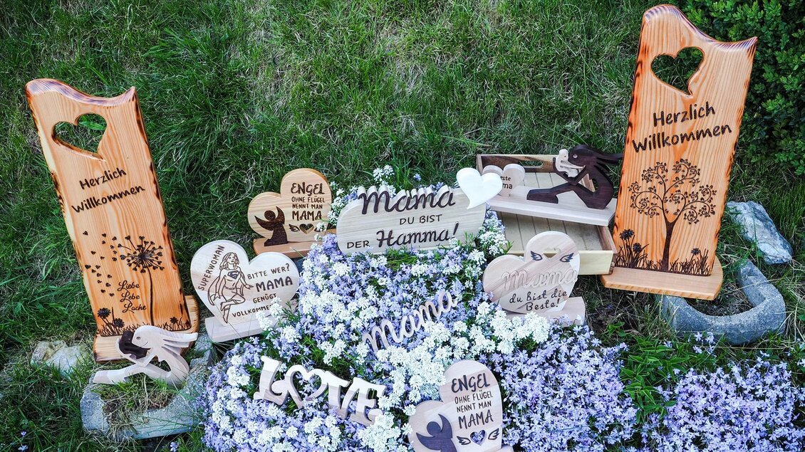 Eine Sammlung von Holzschildern mit einladenden und liebevollen Sprüchen steht auf einer blühenden Wiese. Die Schilder zeigen Botschaften wie "Herzlich Willkommen" und "Mama, du bist der Hammer!". | © Minimi