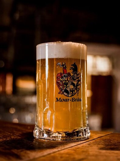 Ein Glas Bier mit cremigem Schaum steht auf einem Holztisch. Im Hintergrund sind verschwommene Lichter und eine gemütliche Atmosphäre zu erkennen. | © Moarpeter