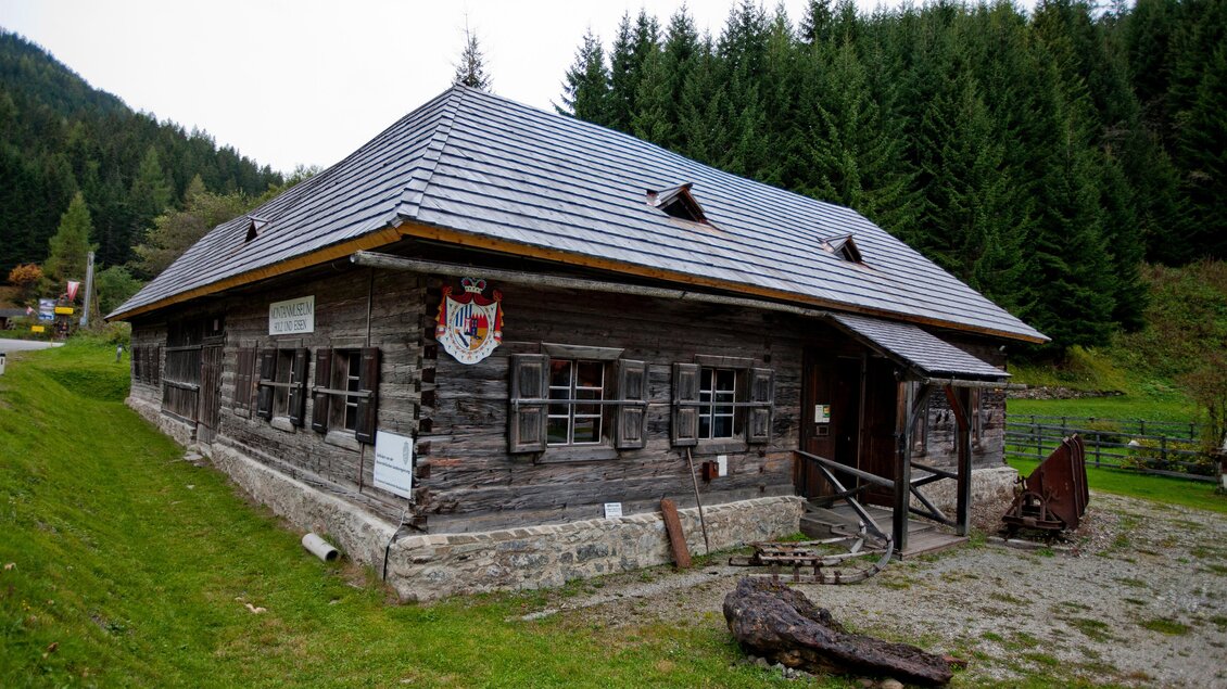 Ein traditionelles Holzhaus mit einem Schrägdach inmitten von grünen Bäumen. Das Gebäude hat kleine Fenster und eine rustikale Fassade. | © Holzwelt Murau