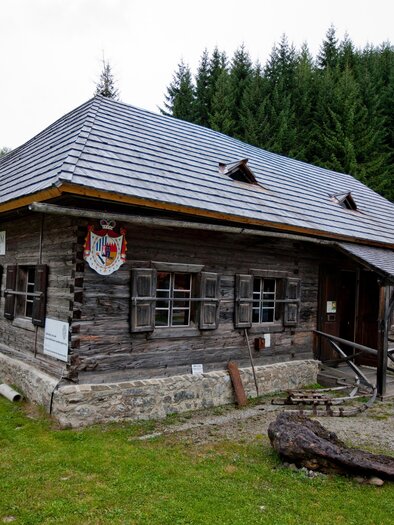 A traditional wooden house with a sloped roof amidst green trees. The building has small windows and a rustic façade. | © Holzwelt Murau