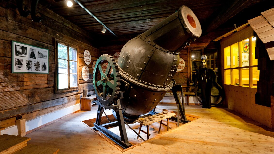 Eine alte Maschine steht in einem rustikalen Raum. Die Wände sind aus Holz und es gibt einige historische Bilder in der Nähe. | © Holzwelt Murau